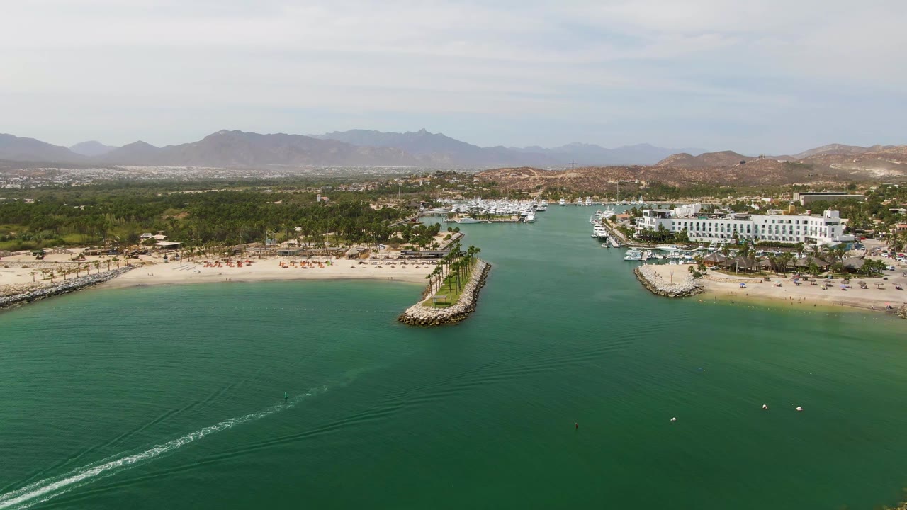 Wide aerial frame over Puerto Los Cabos Beach with turquoise water in front, a faint white boat wake near the lower left, sandy shore, marina boats, white buildings, distant mountains, and no visible people.