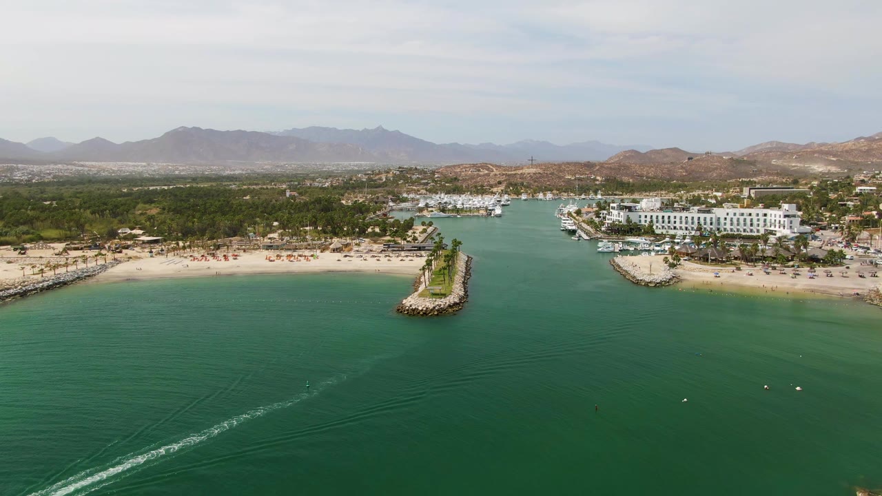 Aerial view at Puerto Los Cabos Beach with the white wake more visible across the lower water, sandy beach and breakwater at midground, marina boats and white buildings beyond, and no visible people.