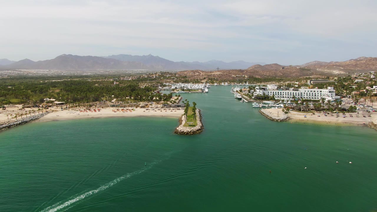 Puerto Los Cabos Beach aerial frame with the wake trailing through the lower left water, a sandy shoreline and small breakwater centered above it, marina slips and white buildings to the right, and no visible people.