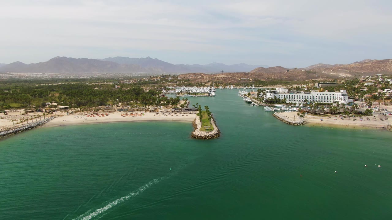 Wide drone frame of Puerto Los Cabos Beach with wake, breakwaters, marina, and mountains