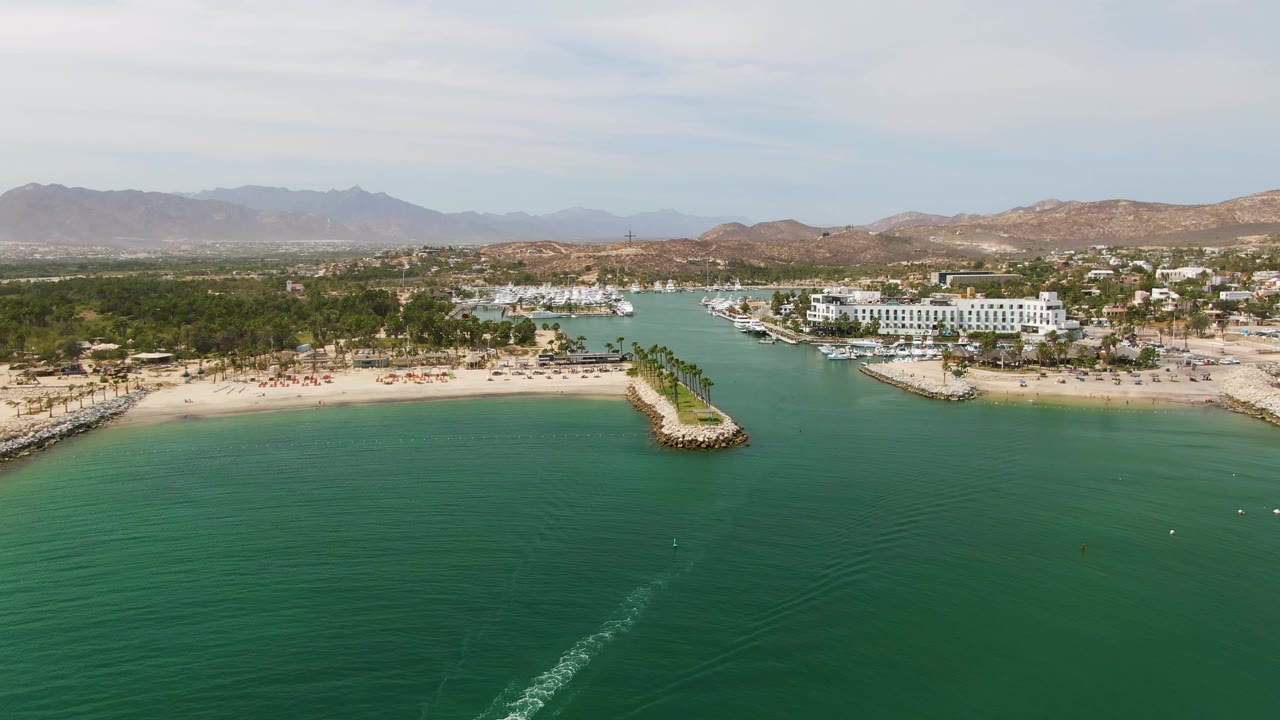 At 0:00, an aerial view over Puerto Los Cabos Beach shows green coastal water, a faint boat wake in the foreground, sandy shoreline, jetties, palm trees, low buildings, distant mountains, and no visible people.