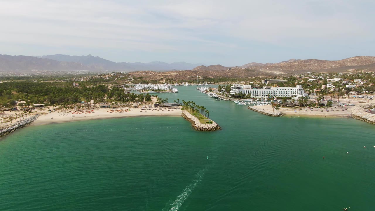 At 0:01, the drone frame holds on Puerto Los Cabos Beach with calm green-blue water, a more central boat wake, sandy beach, rocky breakwaters, palms, shoreline buildings, hills, and no visible people.
