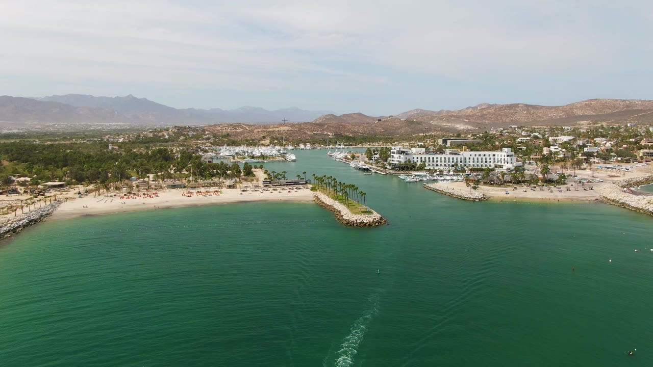 At 0:02, Puerto Los Cabos Beach appears from above with the boat wake trailing through the foreground water, a beach inlet, paired jetties, palms, white shoreline buildings, distant mountains, and no visible people.
