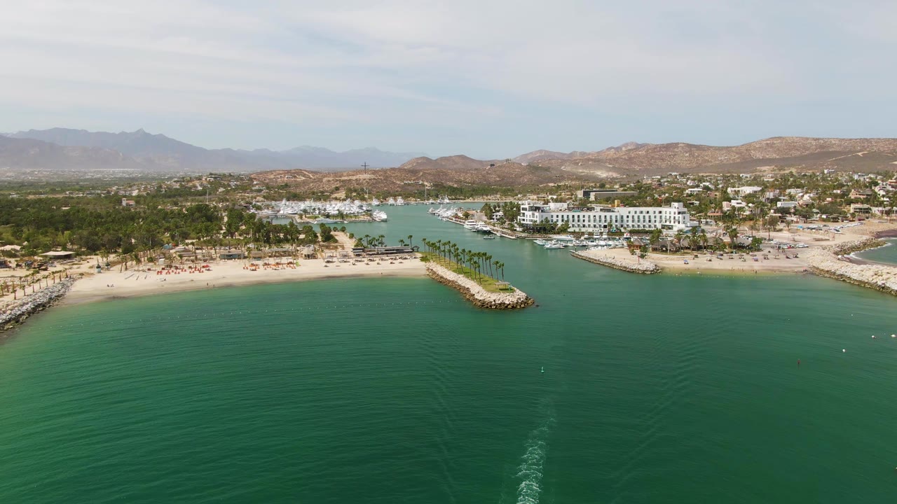 At 0:03, the wide aerial over Puerto Los Cabos Beach shows the wake lower in frame, broad calm water, a palm-lined sandy shore, jetties, coastal buildings, hazy mountains, and no visible people.