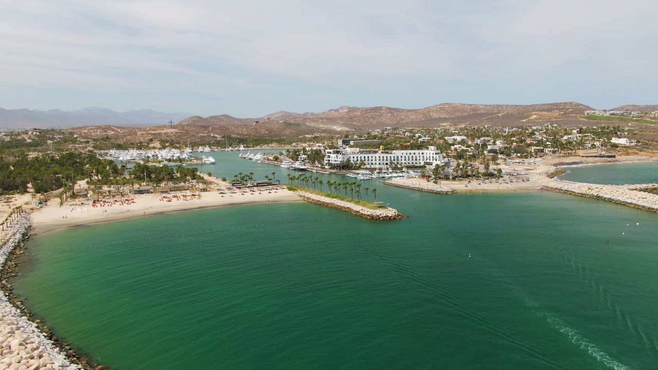 The Puerto Los Cabos Beach shoreline appears in a wide aerial composition, with green-blue water below, a rock breakwater reaching along the sand, palms and white buildings behind it, and no visible people.