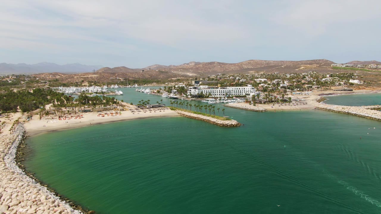 At 0:03, the aerial view shows more of the rock breakwater entering from the lower left, with calm turquoise water beside it, sand and palms across shore, white buildings beyond, and no visible people.