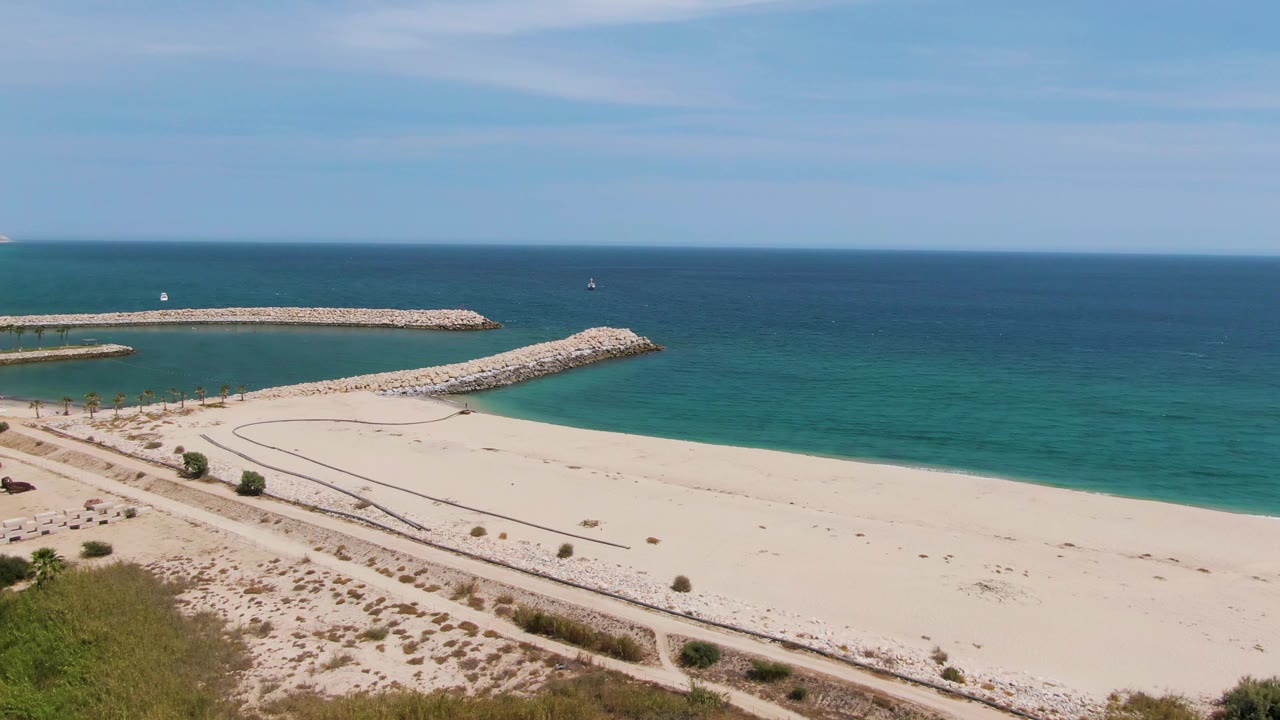 Aerial frame of Puerto Los Cabos Beach with breakwaters offshore, open sand, dune paths, a small access area with vehicles, and people visible near the left side of the beach.