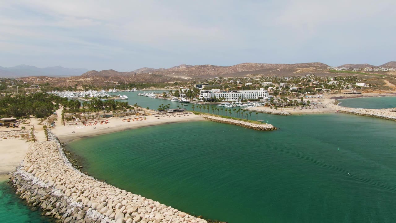 Aerial view of Puerto Los Cabos Beach with a rocky breakwater angling from the lower left toward a sandy beach, turquoise water, palm trees, boats, white buildings, hills, and no visible people.