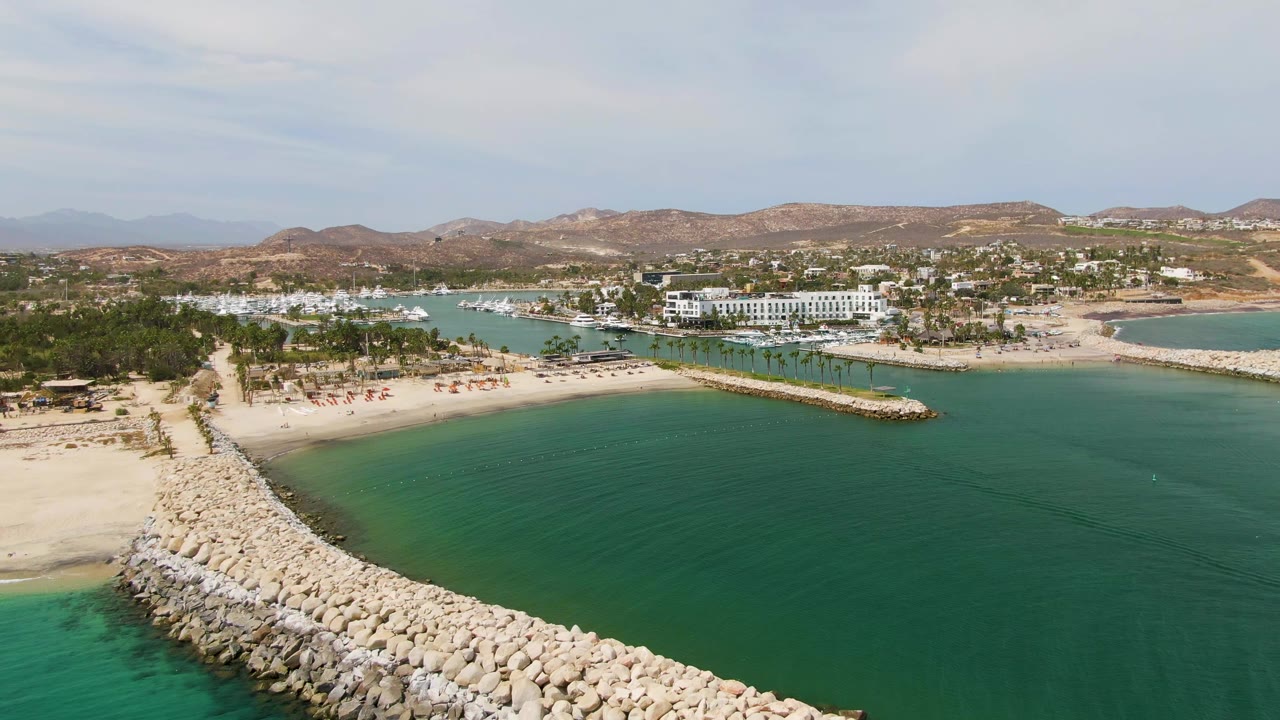Aerial frame at Puerto Los Cabos Beach showing the rocky breakwater more centered in turquoise water, with sandy shore, palm-lined beach area, boats, low buildings, dry hills, and no visible people.