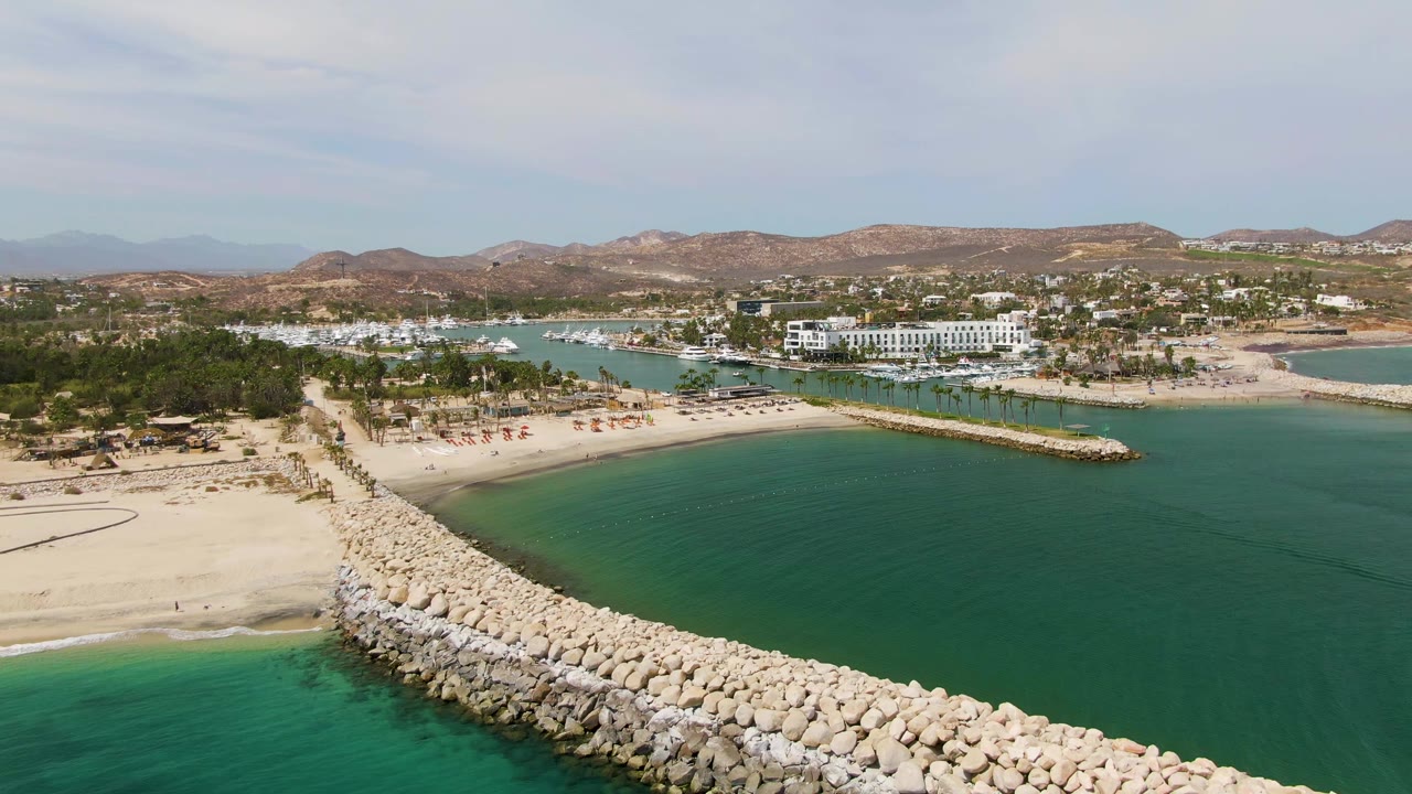 Sandy beach and rocky breakwater seen from above with turquoise water and hills beyond.