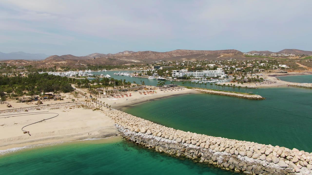 Aerial frame over Puerto Los Cabos Beach showing a pale rock breakwater curving across turquoise water, sandy shore, palms, resort buildings, low hills, clouded sky, and no visible people.
