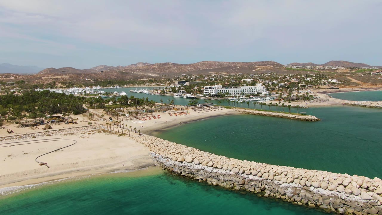 Puerto Los Cabos Beach aerial view with the rock breakwater spanning the lower frame, green-blue ocean in front, pale sand and palms behind it, distant resort buildings, hills, and no visible people.