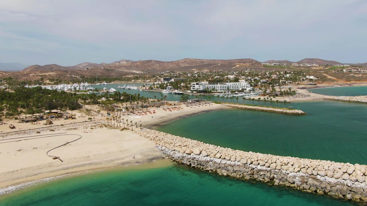 Drone frame of Puerto Los Cabos Beach with the rocky breakwater crossing the foreground, a small sandy beach and turquoise bay behind it, white resort buildings, brown hills, light clouds, and no visible people.