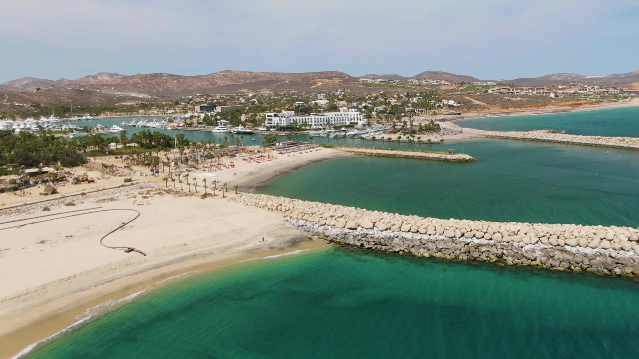 Puerto Los Cabos Beach aerial frame with turquoise water in the foreground, a pale sandy curve, a rock breakwater, harbor boats, white buildings, mountains, and no visible people.