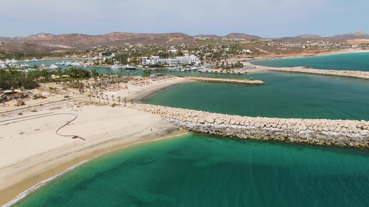 At 0:01, the Puerto Los Cabos Beach shoreline fills the lower frame with clearer green shallows, the rock breakwater spans the center, harbor boats sit behind it, and no visible people appear.