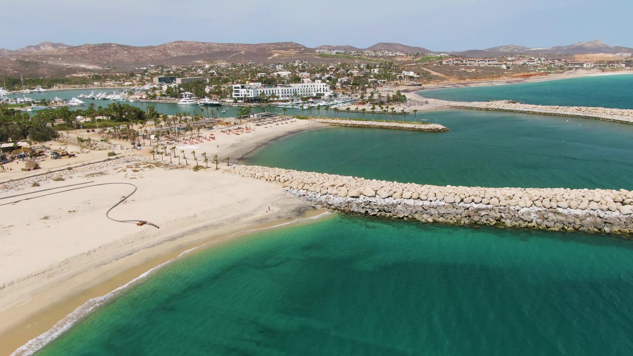 At 0:02, Puerto Los Cabos Beach is seen from above with darker underwater patches in the foreground water, a broad sandy beach, a long rock breakwater, distant harbor buildings, and no visible people.