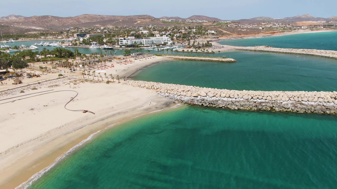 Aerial coastal view with sandy shoreline, gentle surf, rock breakwater, harbor, buildings, palms, and mountains.