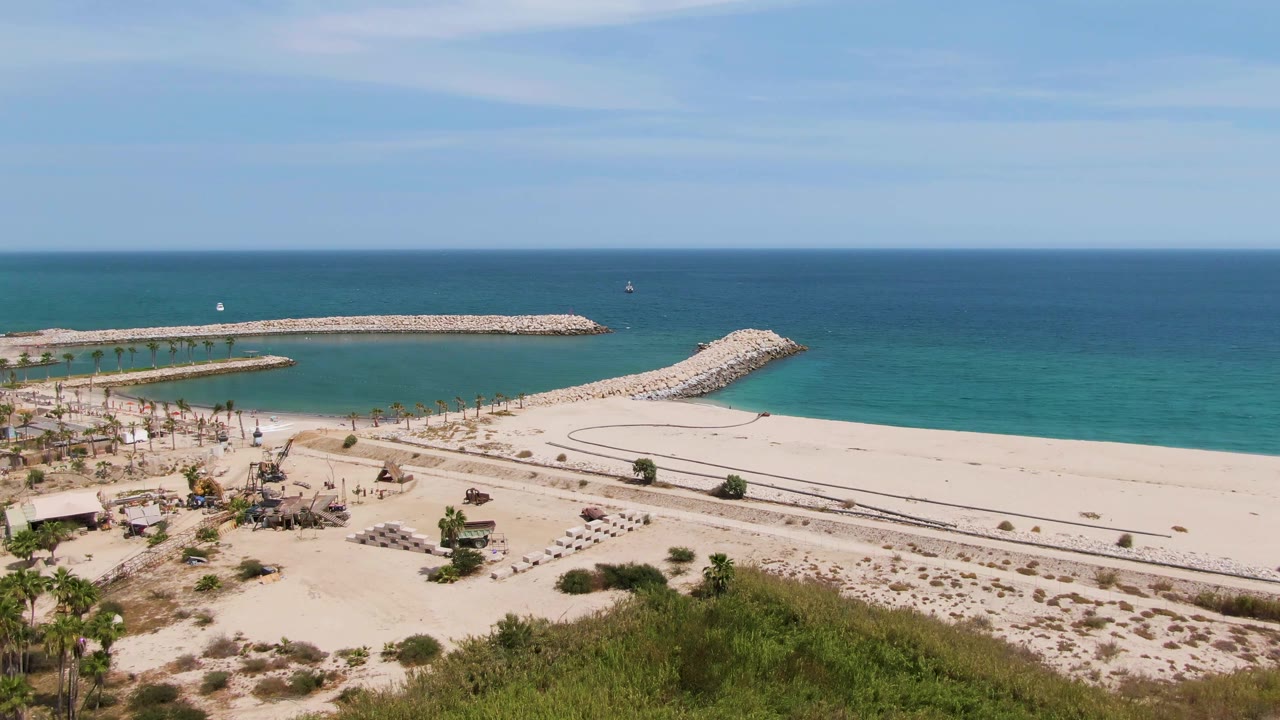 Wide aerial view over Puerto Los Cabos Beach with pale sand, foreground palms, curving rock breakwaters, turquoise shallows, deep blue ocean, light cloud streaks, and no visible people.