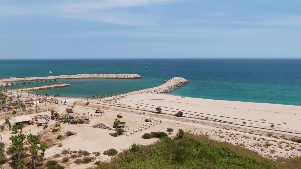 Puerto Los Cabos Beach from above with the long rock jetty stretching across the water, a shorter breakwater angling from the sand, palm landscaping at lower left, and no visible people.