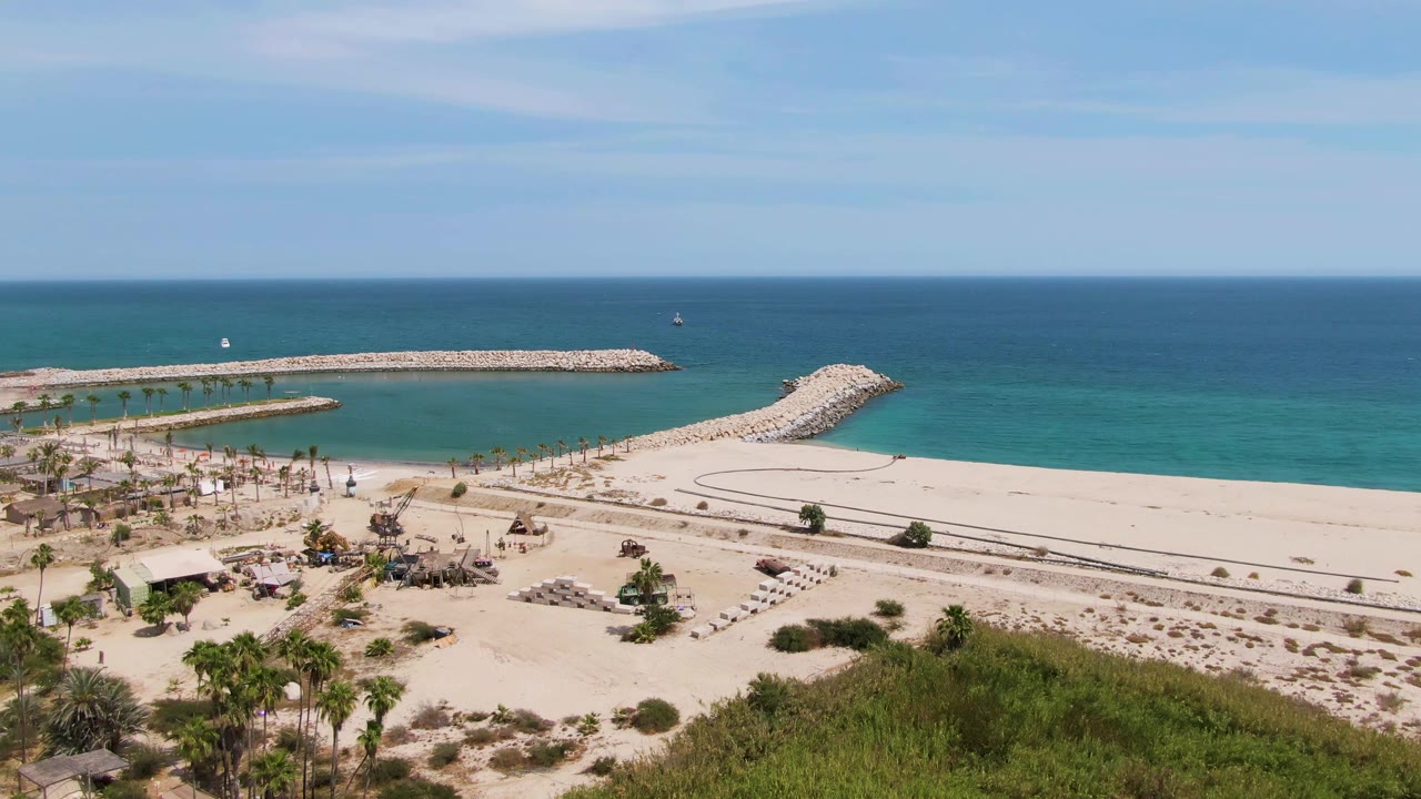 Aerial coastal scene with palms, beach paths, breakwaters, and blue water.