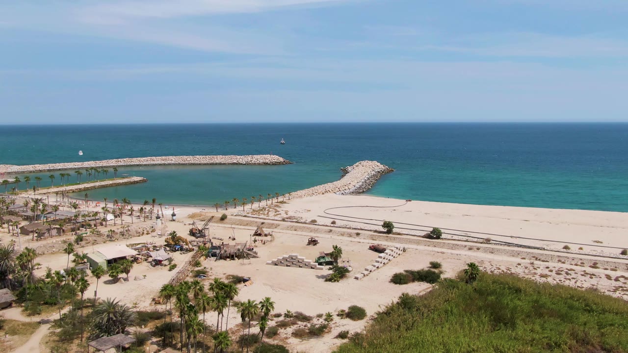 Aerial view over Puerto Los Cabos Beach showing pale sand, palm trees, lounge chairs, stone breakwaters, turquoise water, and no clearly visible people at the start of the clip.