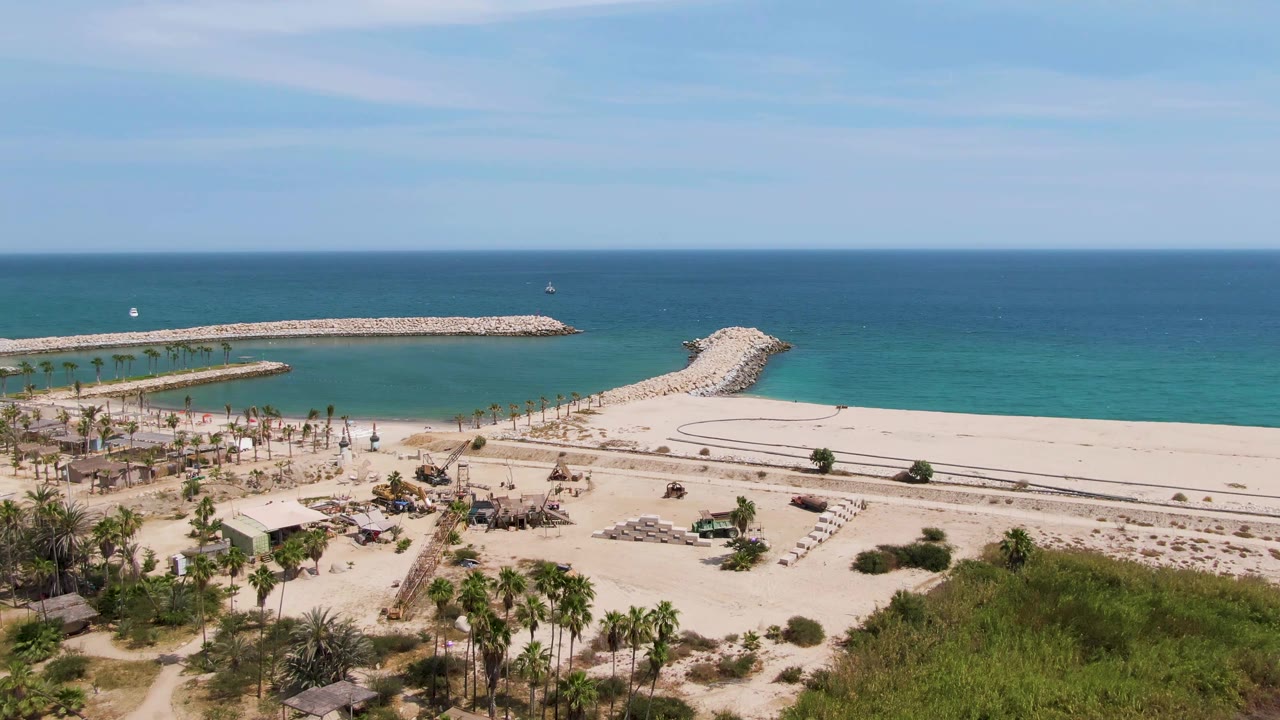 Puerto Los Cabos Beach appears from above with paired rock breakwaters extending into clear water, palms and lounge chairs on the sand, low green vegetation, and no clearly visible people.