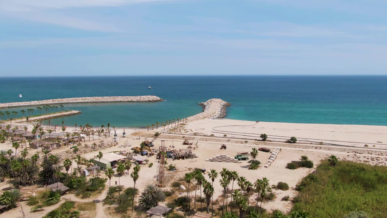 Opening aerial frame over Puerto Los Cabos Beach with palms and huts in the foreground, a sandy beach road, rocky breakwaters, blue ocean, and no visible people.