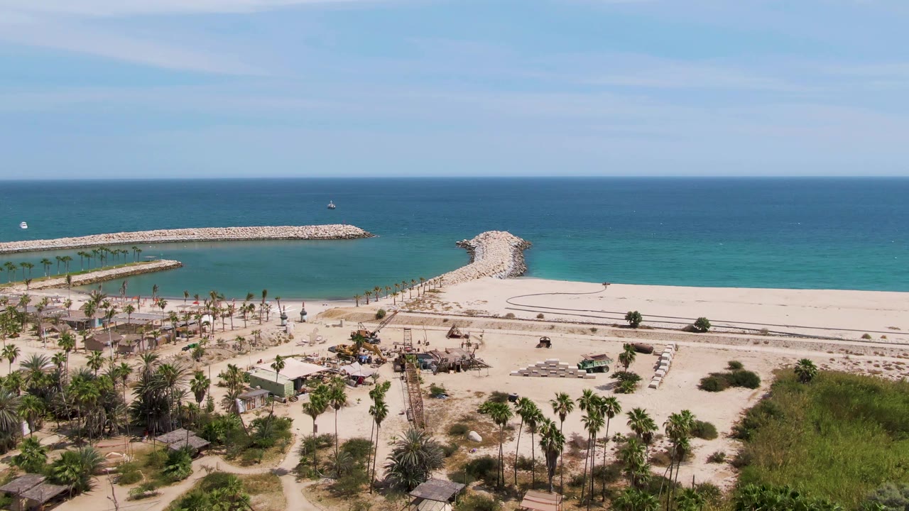 Puerto Los Cabos Beach frame at two seconds with the palm cluster and huts low in frame, pale sand and road across the middle, and blue ocean beyond the breakwaters, with no visible people.