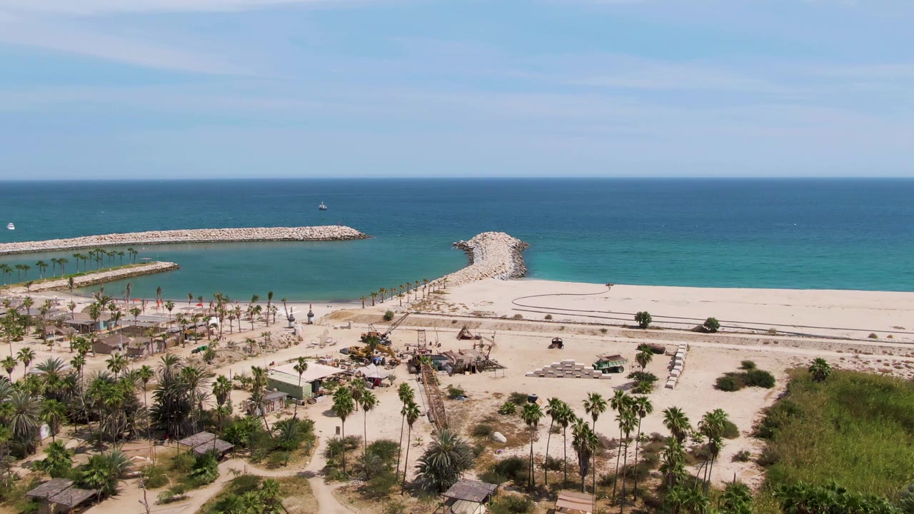 Drone-height view of beach huts, palm trees, sand, breakwaters, and blue sea.
