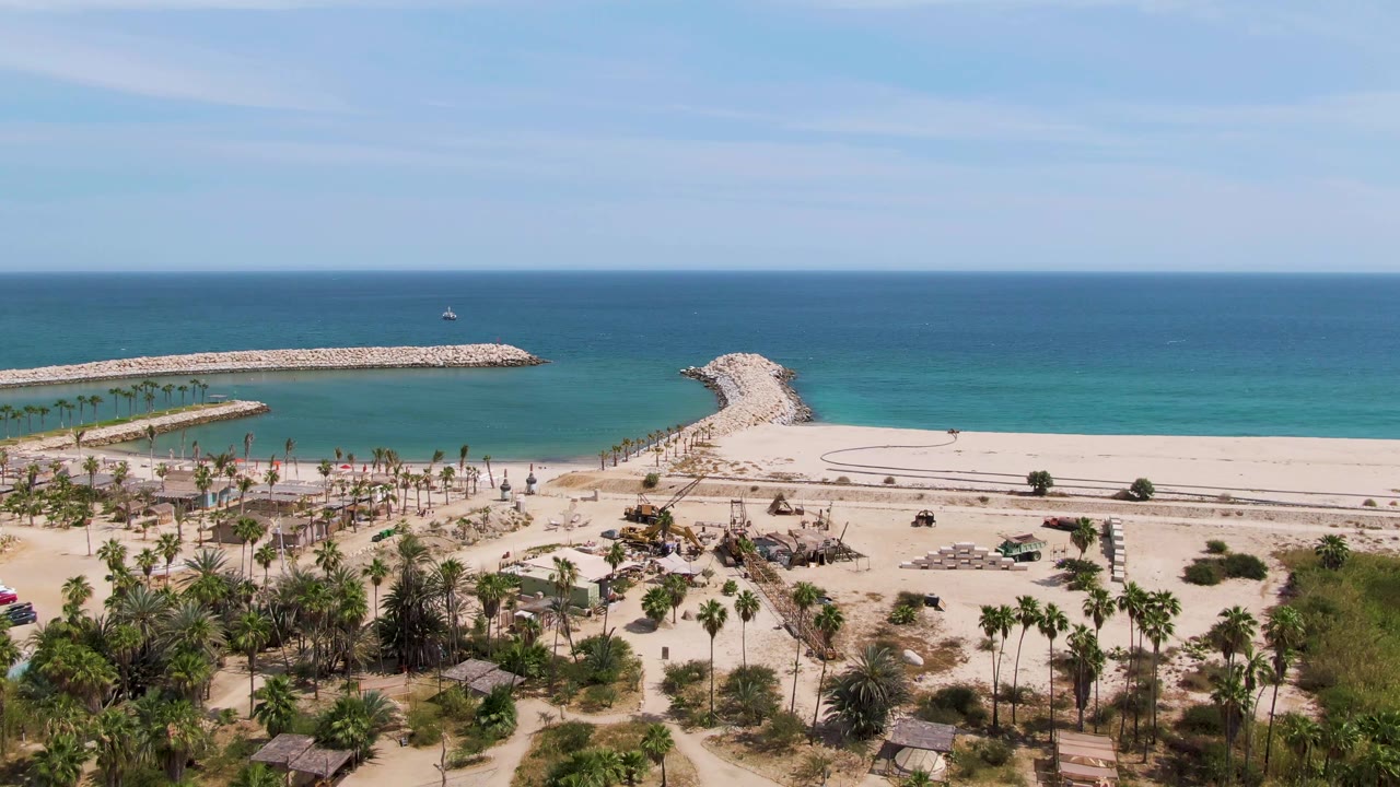 At 0:01, the aerial view of Puerto Los Cabos Beach shows small cars along the coastal road, palms below, pale sand, turquoise shallows, a rocky breakwater, and no visible people.