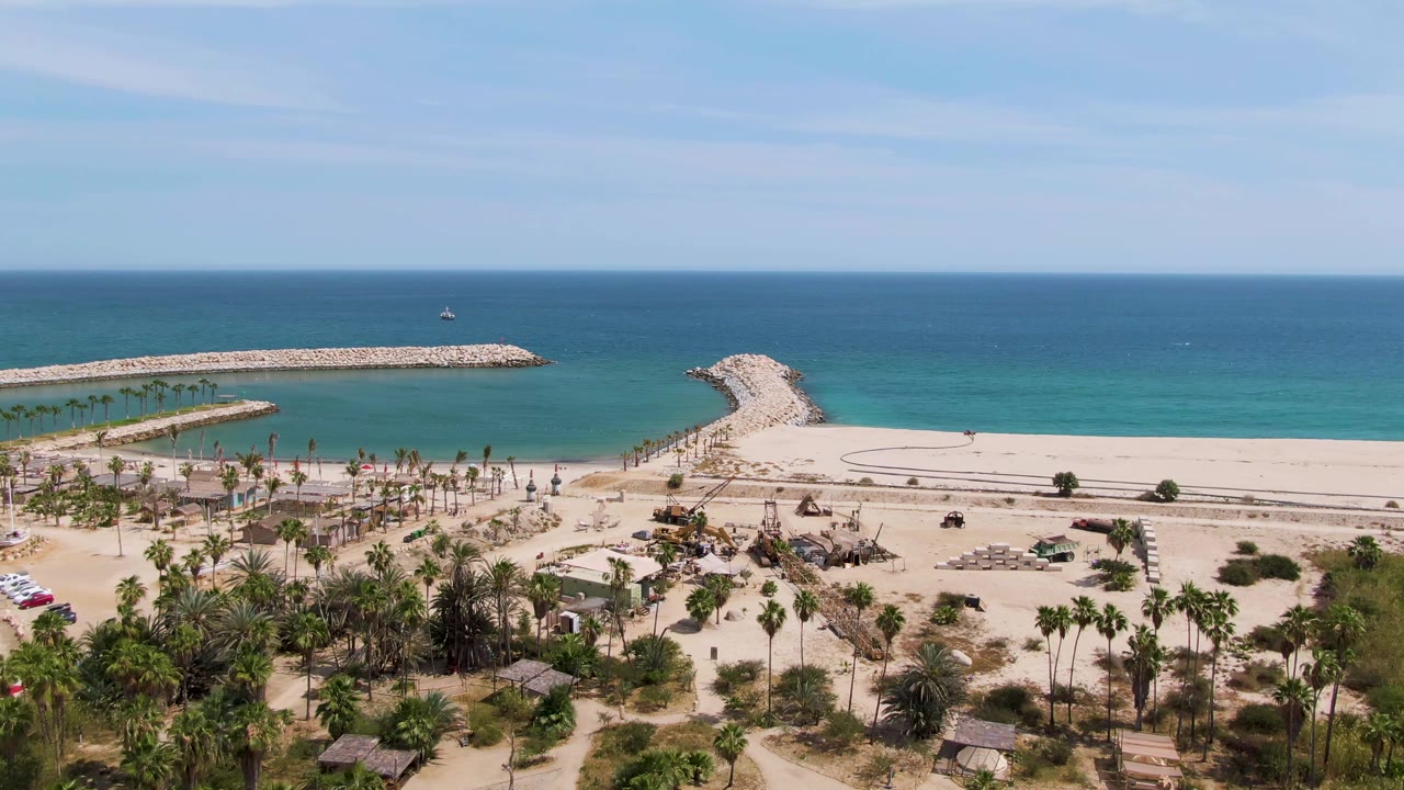 At 0:02, Puerto Los Cabos Beach is framed with the palm grove and beach road low in the image, the curved breakwater near center, deep blue ocean behind it, and no visible people.