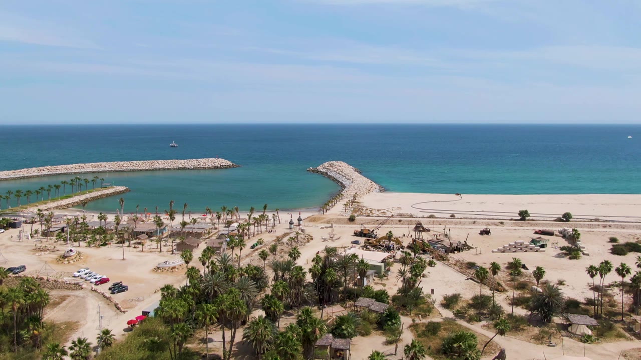 Puerto Los Cabos Beach appears from above with the two stone breakwaters spanning the shoreline, a palm-lined sandy area below, turquoise water under a clear sky, and no visible people.