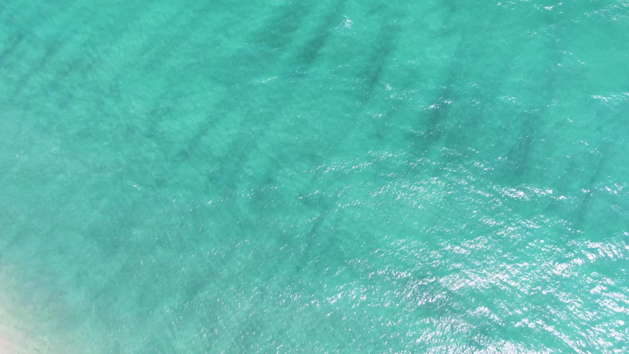 Overhead view of clear turquoise ocean water at Puerto Los Cabos Beach with soft ripples, faint darker bands below the surface, and no visible people.