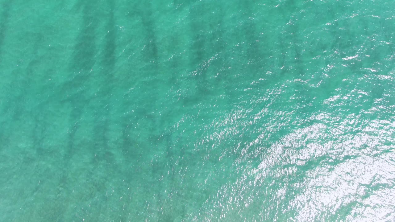 Overhead turquoise water at Puerto Los Cabos Beach with a concentrated patch of white surface reflection on the right and no visible people.