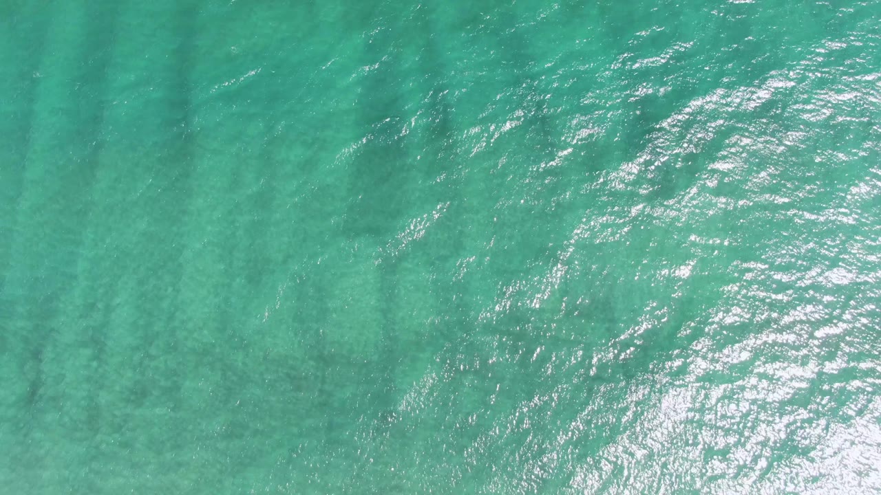 Overhead frame of clear turquoise sea at Puerto Los Cabos Beach, showing long dark underwater bands, small wind ripples, scattered light flecks, and no visible people.