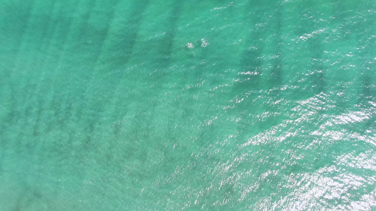 Top-down Puerto Los Cabos Beach water frame with turquoise ripples, a broad bright sunlight reflection near the lower left, faint dark seabed streaks, and no visible people.
