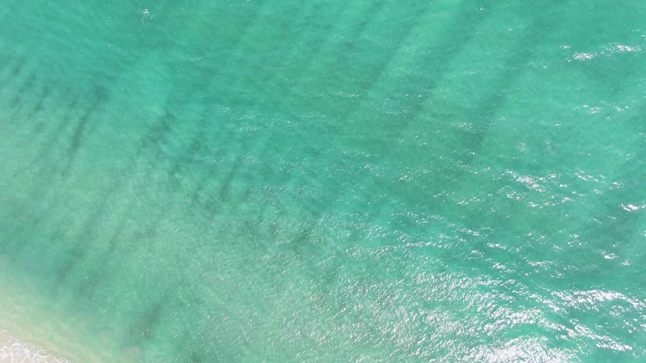 Overhead view of turquoise water and pale sand at Puerto Los Cabos Beach, with faint foam at the lower edge and no visible people.