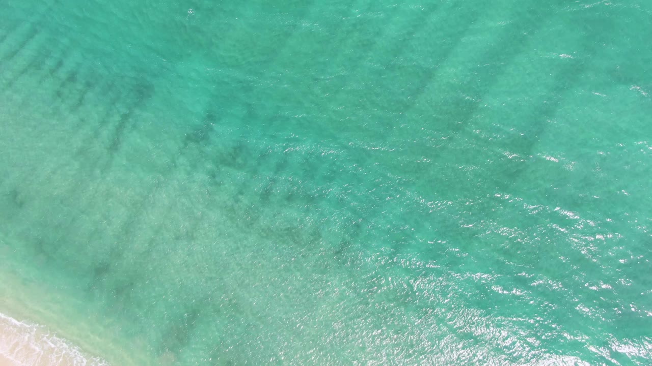 Puerto Los Cabos Beach seen from above as green-blue water ripples over shallow sand, with a narrow white surf line and no visible people.