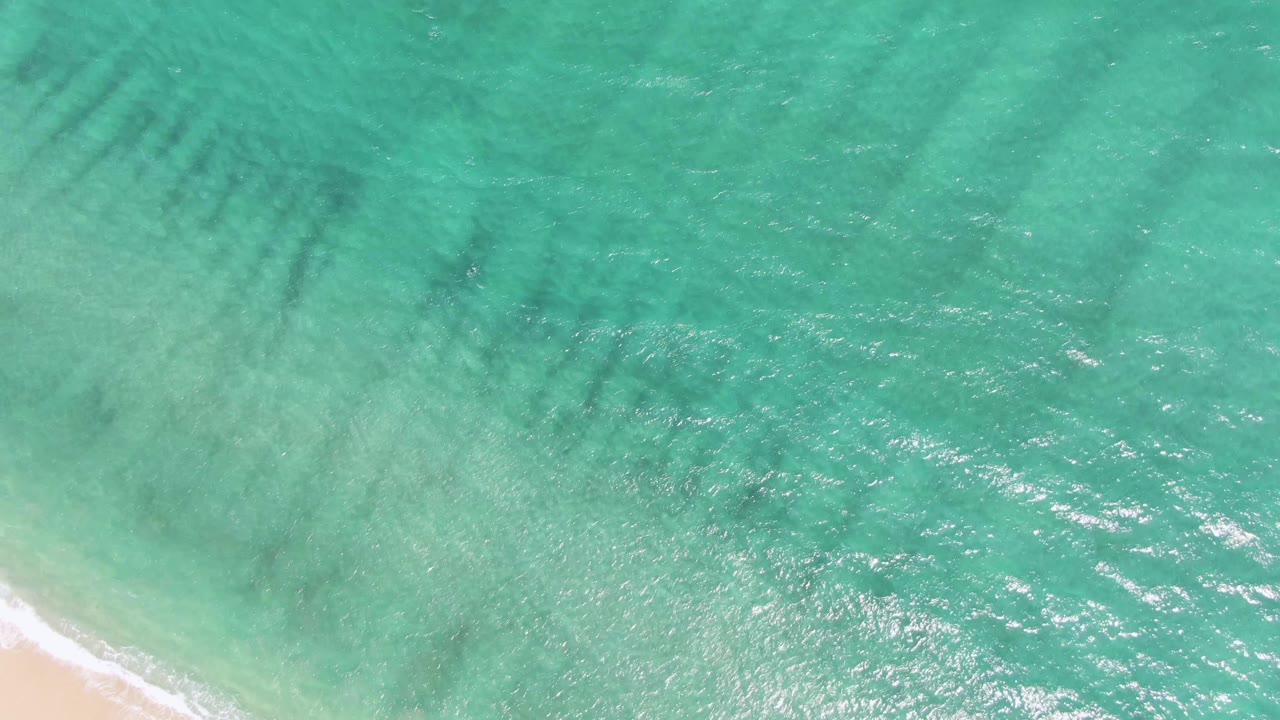 Aerial frame over Puerto Los Cabos Beach with turquoise water filling the view, darker underwater streaks near the top, bright surface glints, and no visible people.