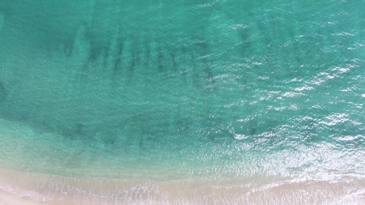 Top-down view at Puerto Los Cabos Beach with bright turquoise surf, darker submerged bands in the upper water, and no visible people.