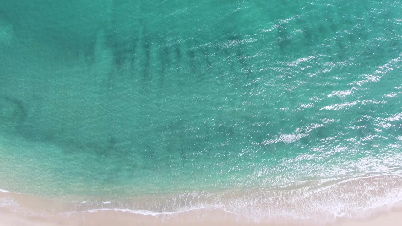 Overhead Puerto Los Cabos Beach shoreline frame with turquoise water filling most of the view, foam spreading along the sand, and no visible people.