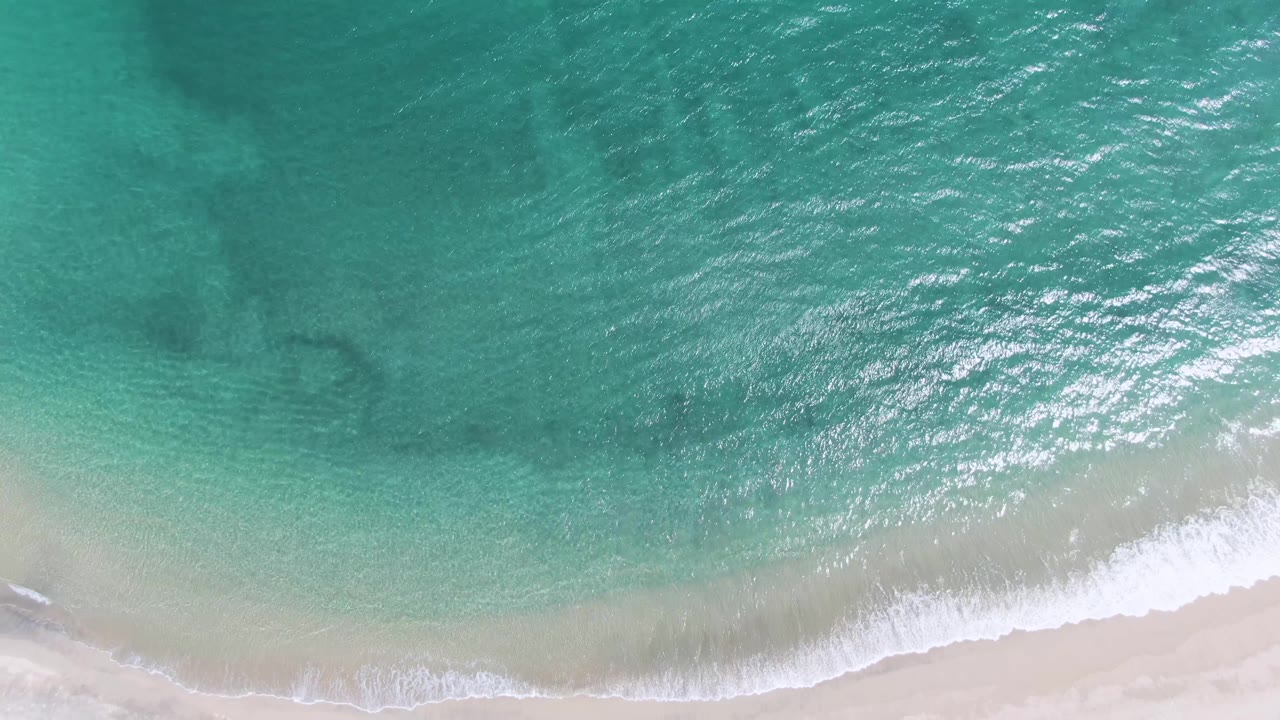 Aerial frame of Puerto Los Cabos Beach where the foam line curves across the lower edge and sunlight catches ripples in the turquoise water, with no visible people.