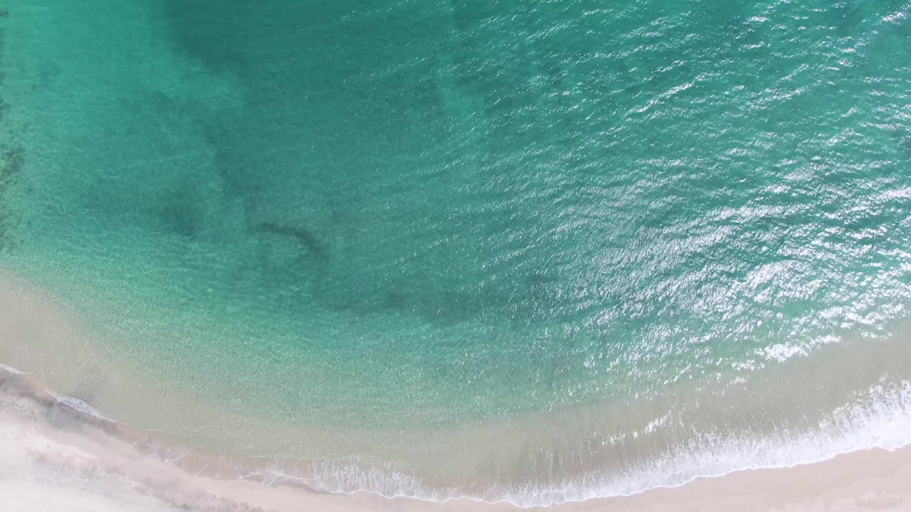 Overhead Puerto Los Cabos Beach frame with a thicker band of white foam near the lower center, pale sand below it, and textured turquoise water above, with no visible people.