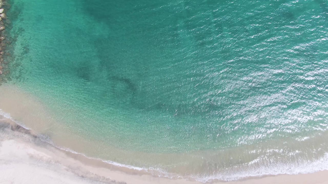 Top-down frame of Puerto Los Cabos Beach with the shoreline set diagonally along the lower part of the image, a light sandy shallows band, and no visible people.