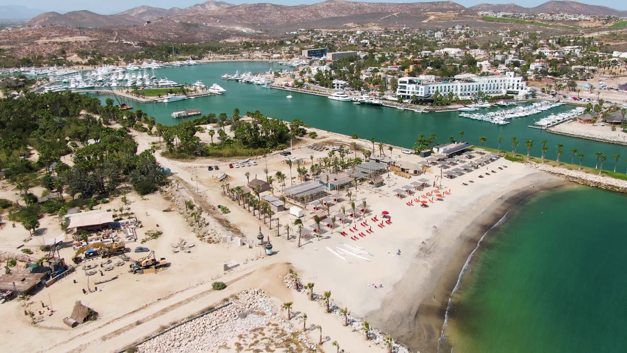 Aerial frame over Puerto Los Cabos Beach showing sand, turquoise shoreline water, red lounge chairs, construction equipment, palm trees, marina docks, hills, and no visible people.