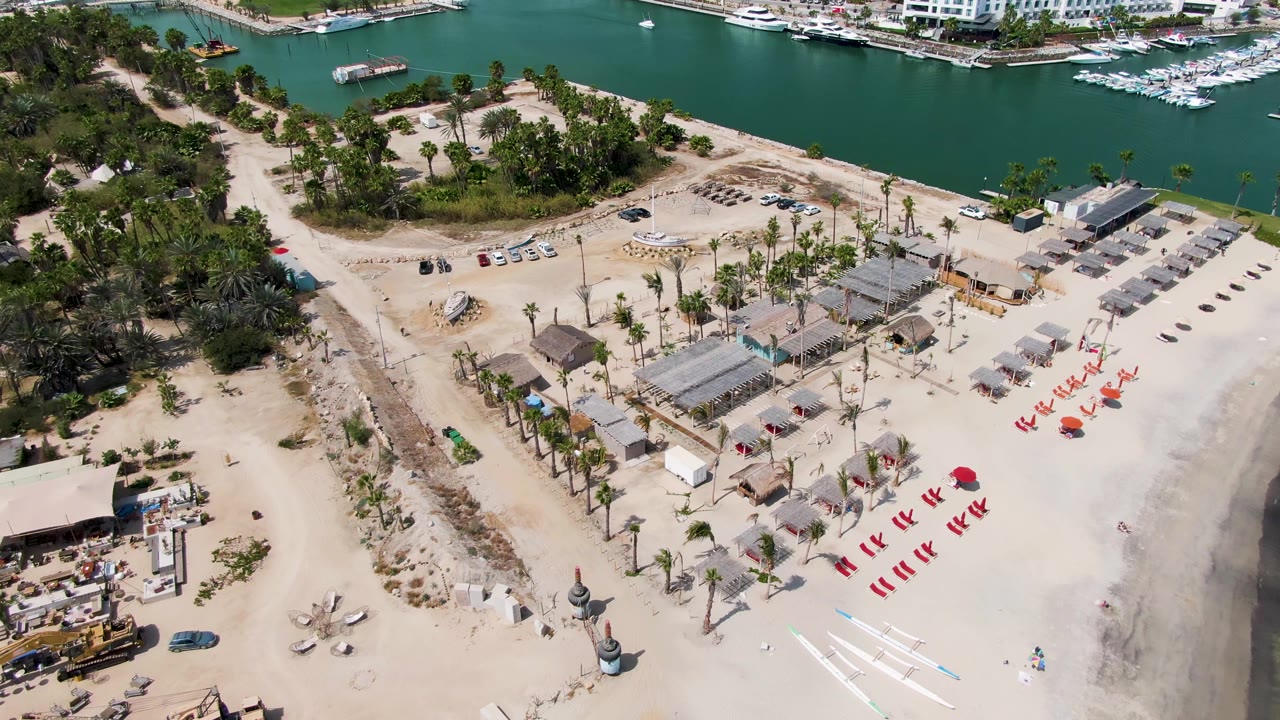 High aerial frame of Puerto Los Cabos Beach with palm-lined sand, shaded huts, red chairs, construction zones, green-blue marina water, boats, and no visible people.