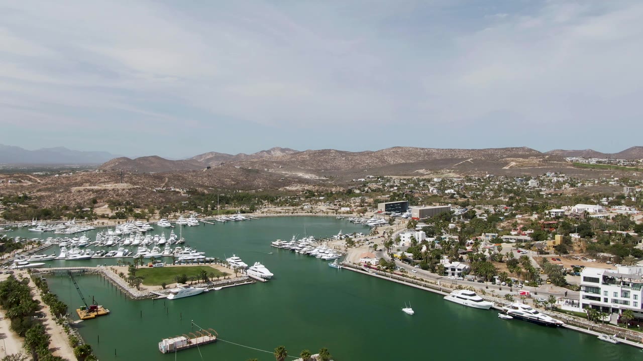 High aerial frame over Puerto Los Cabos Beach marina with green water in front, docked white yachts along the shoreline, dry hills beyond, and no visible people.