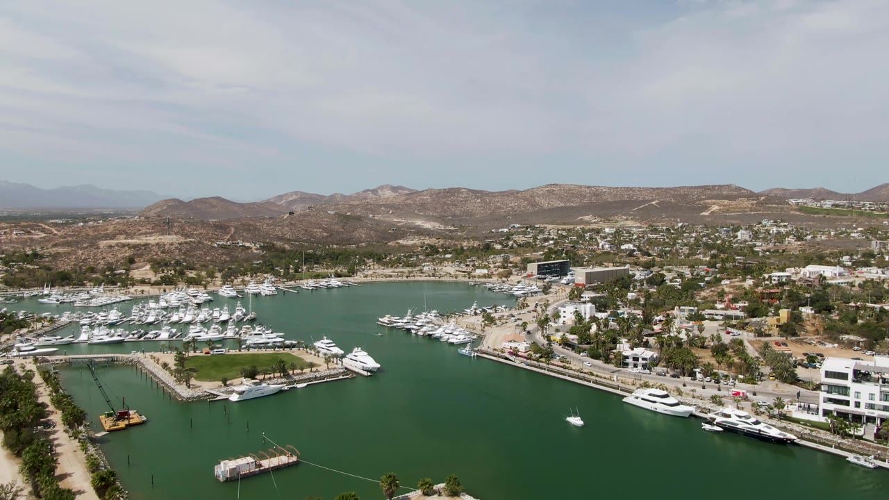 Puerto Los Cabos Beach marina seen from above with a waterfront road near the center, rows of docked yachts, green harbor water, hillside terrain, and no visible people.