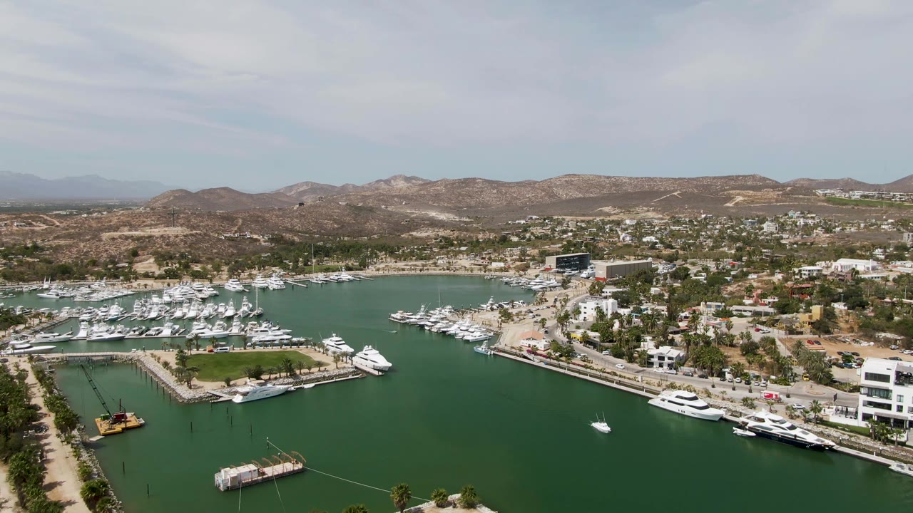 Wide Puerto Los Cabos Beach marina frame with a small white boat crossing open green water, yacht docks on both shorelines, low buildings, dry hills, and no visible people.