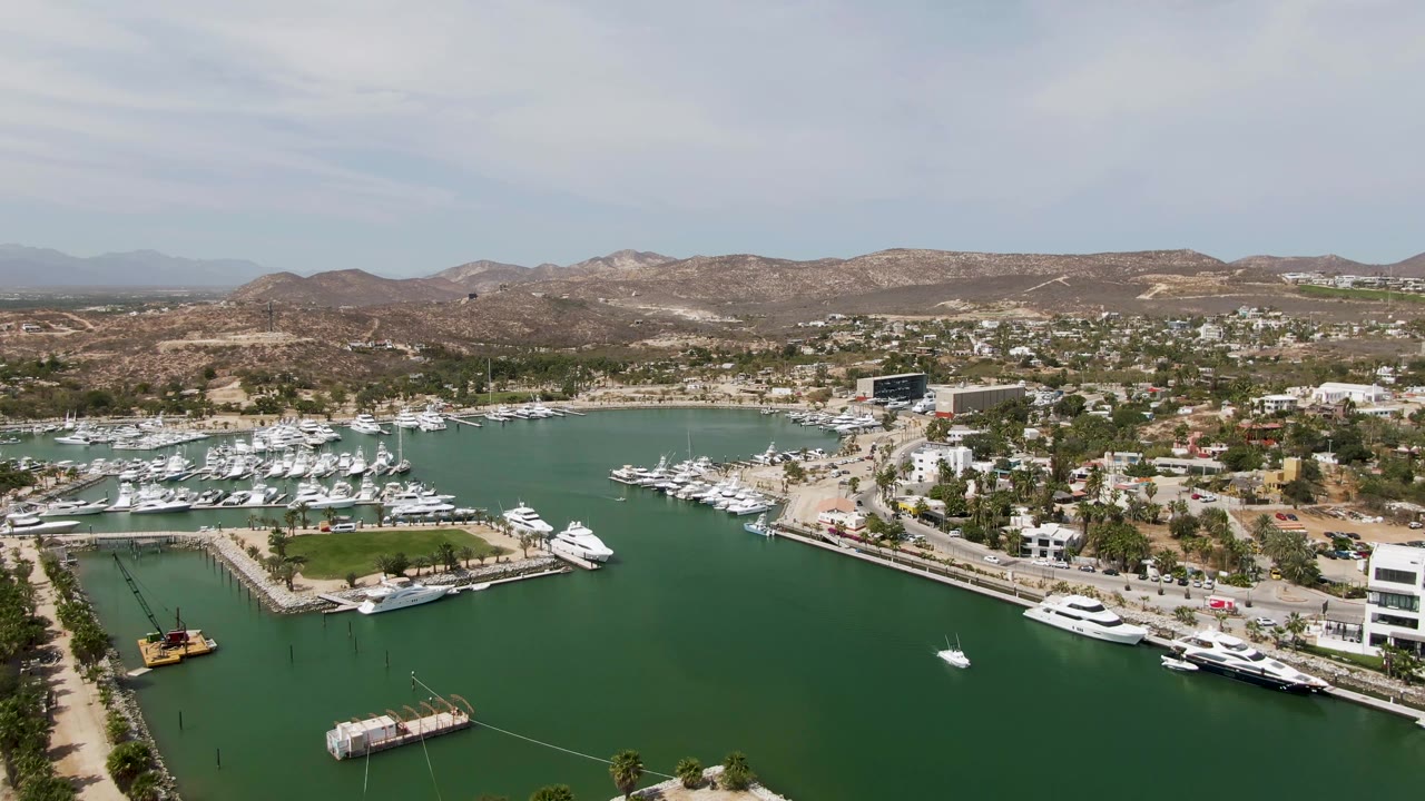 Aerial view of marina slips, yachts, green water, buildings, and low brown hills.